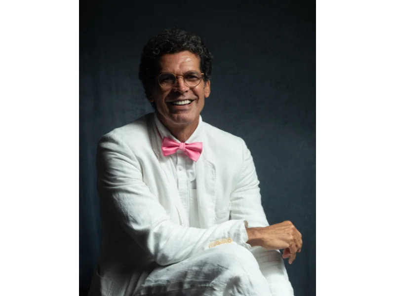 Jackson Costa, Ator e Diretor, está sentado com braço apoiado na perna, vestido conjunto de terno branco e gravata tipo borboleta na cor rosa.
