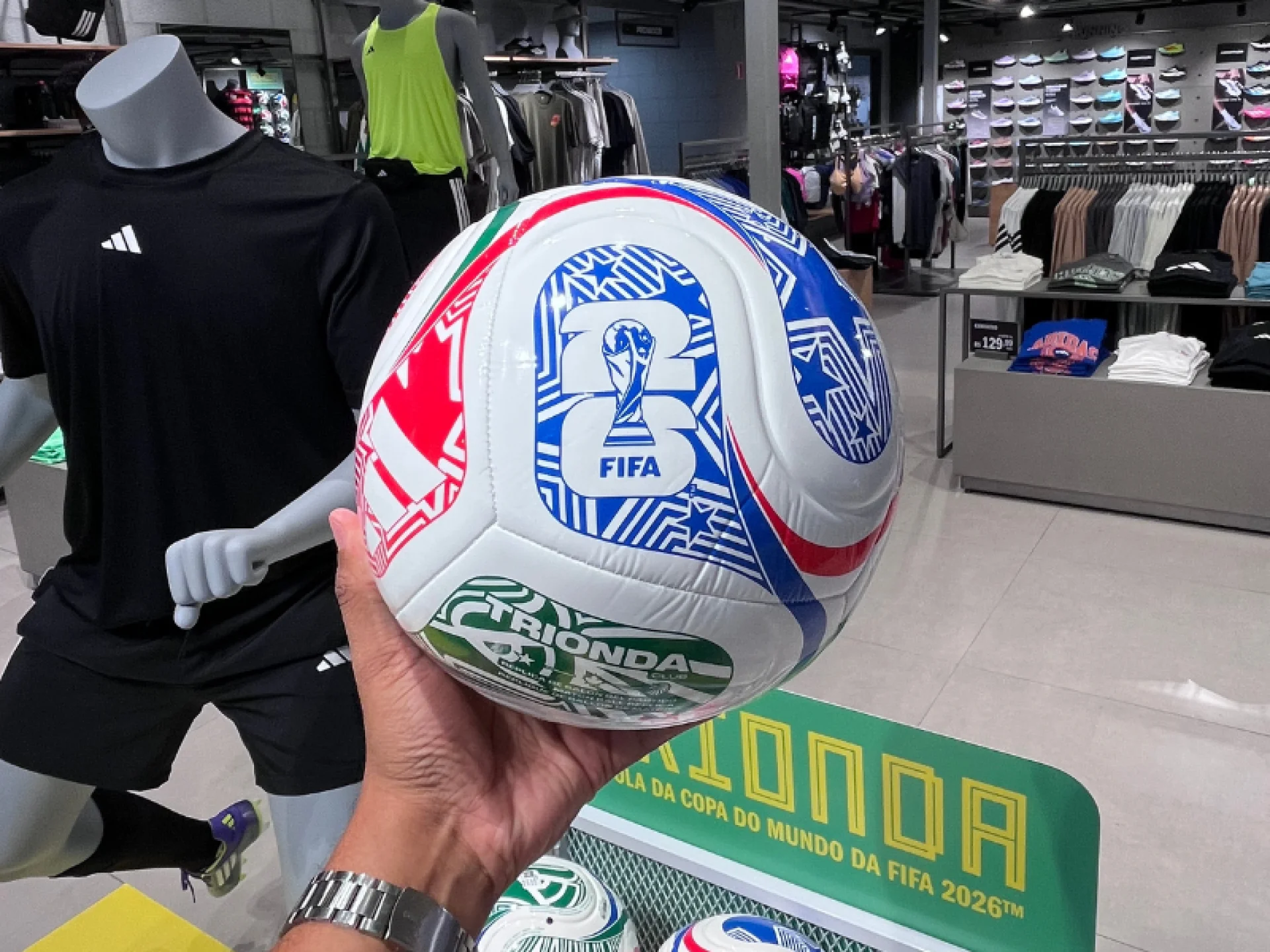 Mão masculina segurando a bola Trionda com a parte da taça da FIFA virada para câmera; Ao lado um manequim com short e camisa da cor preta, também da marca adidas.