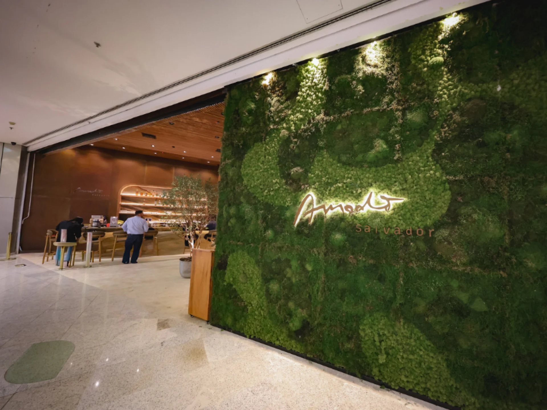 Fachada do restaurante Amado, com uma parede composta de vegetação e o letreiro do nome do restaurante centralizado na parede com LED iluminando.