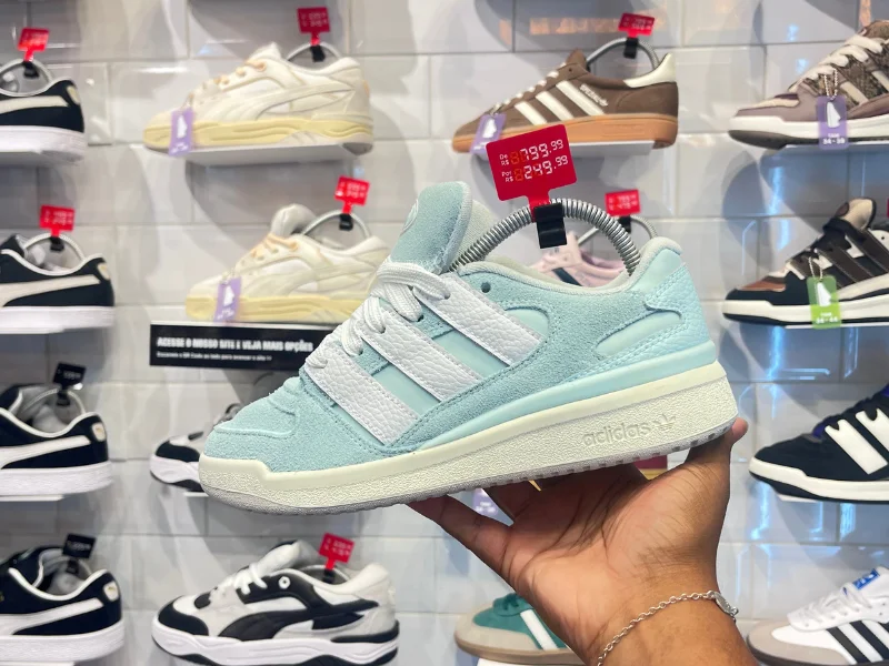 Mão masculina segurando tênis da marca Adidas na cor azul clarinho com símbolo da marca na cor branca. No tênis também tem a etiqueta de promoção de R$799,99 por R$249,99