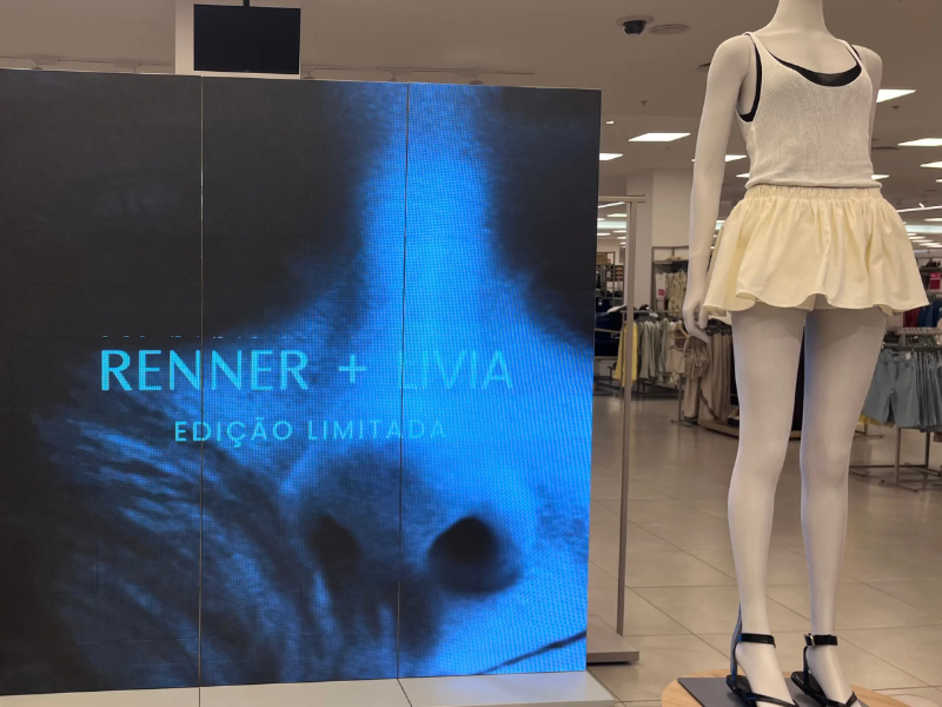 LED com foto da Lívia com letreiro "Renner + Lívia - Edição limitada" e manequim com uma mini-saia cor off-white e regata da cor preta com outra regata de cor branca, peças que fazem parte da coleção.
