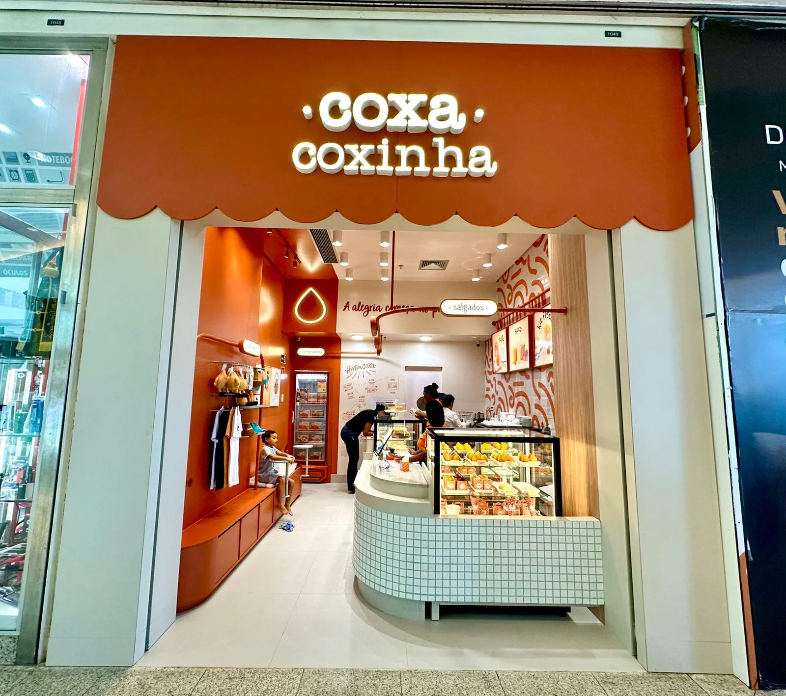 Parte externa da loja com testeira na cor laranja e letreiro com o nome da loja "coxinha coxinha" em caixaria e iluminado, paredes laterais na cor creme